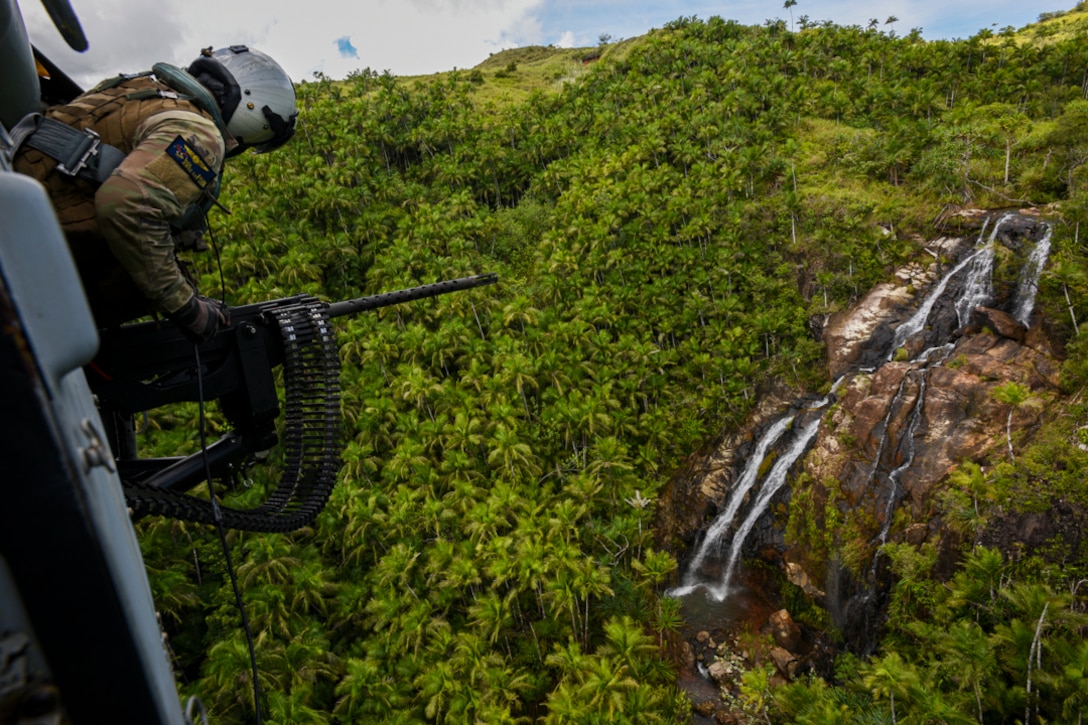 A sailor in a flight helmet looks out from a flying helicopter toward a mountain waterfall.
