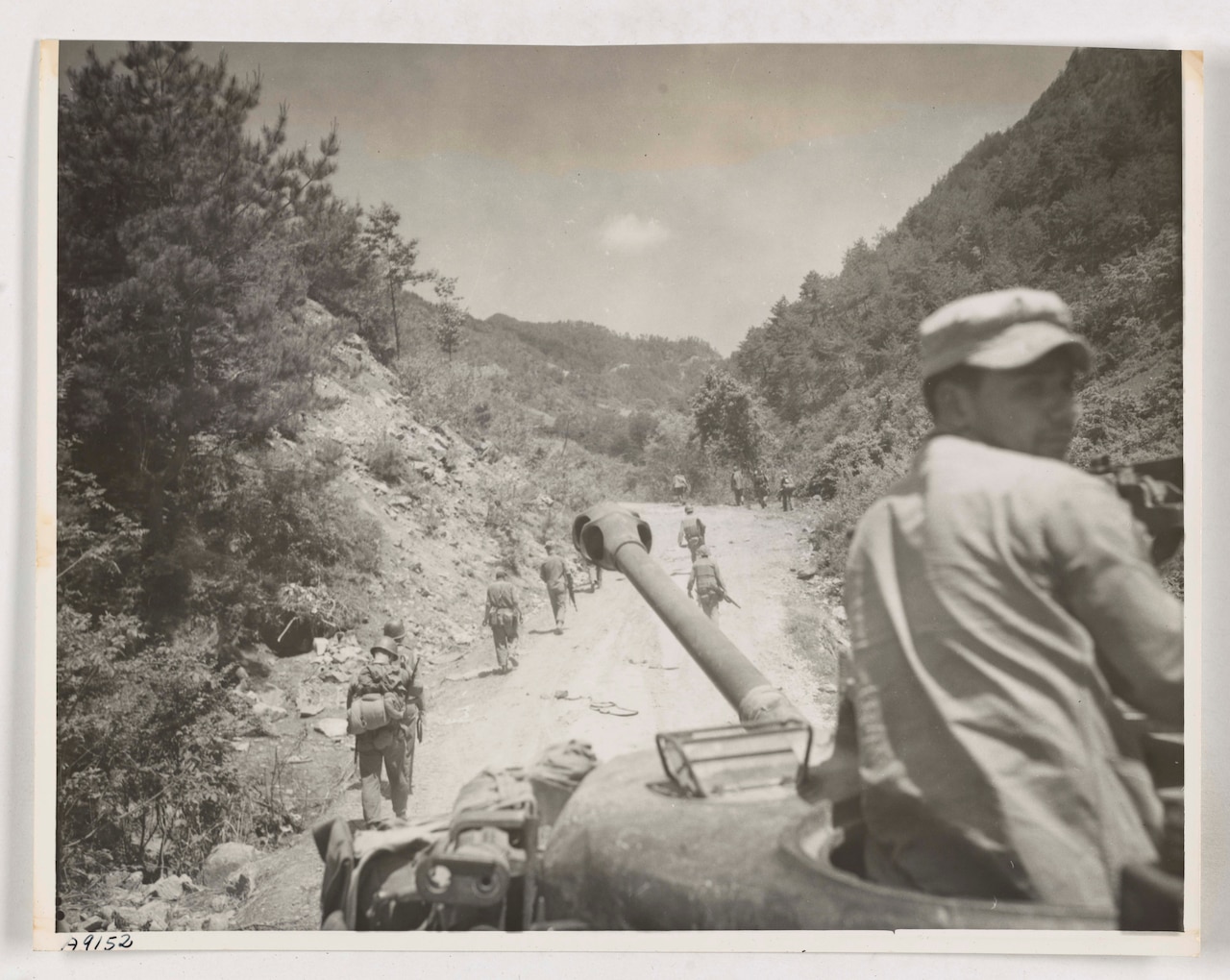 A man emerging from the top of a tank looks over his shoulder. Marines walk on the road ahead of the tank.