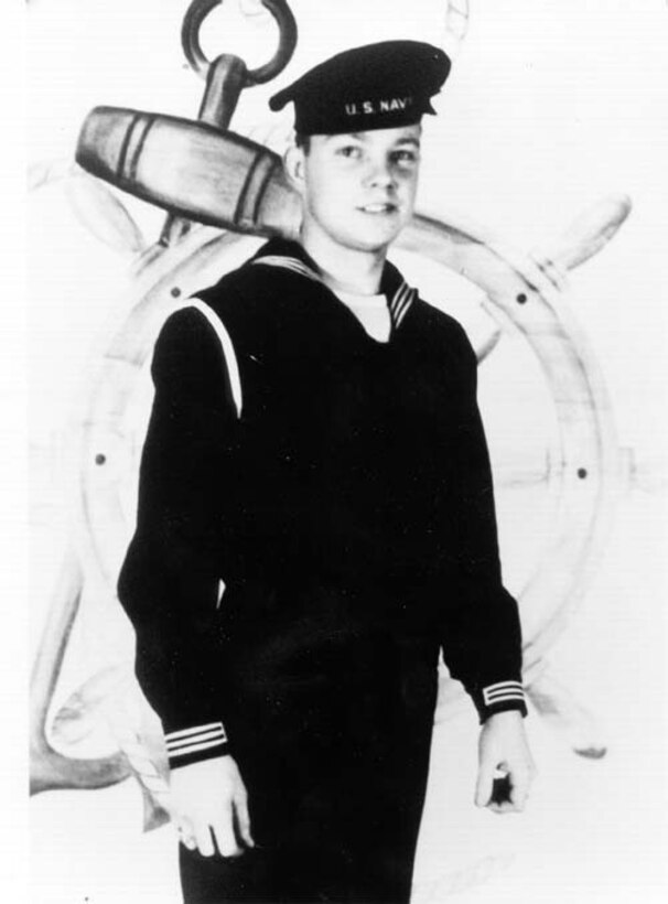 A young sailor in a Navy uniform and flat-top cap poses for a photo.