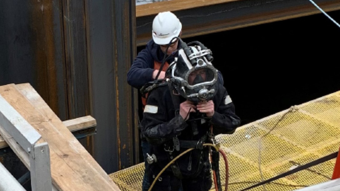 A man is in a diving suit, another man in a hard hat stands behind him.