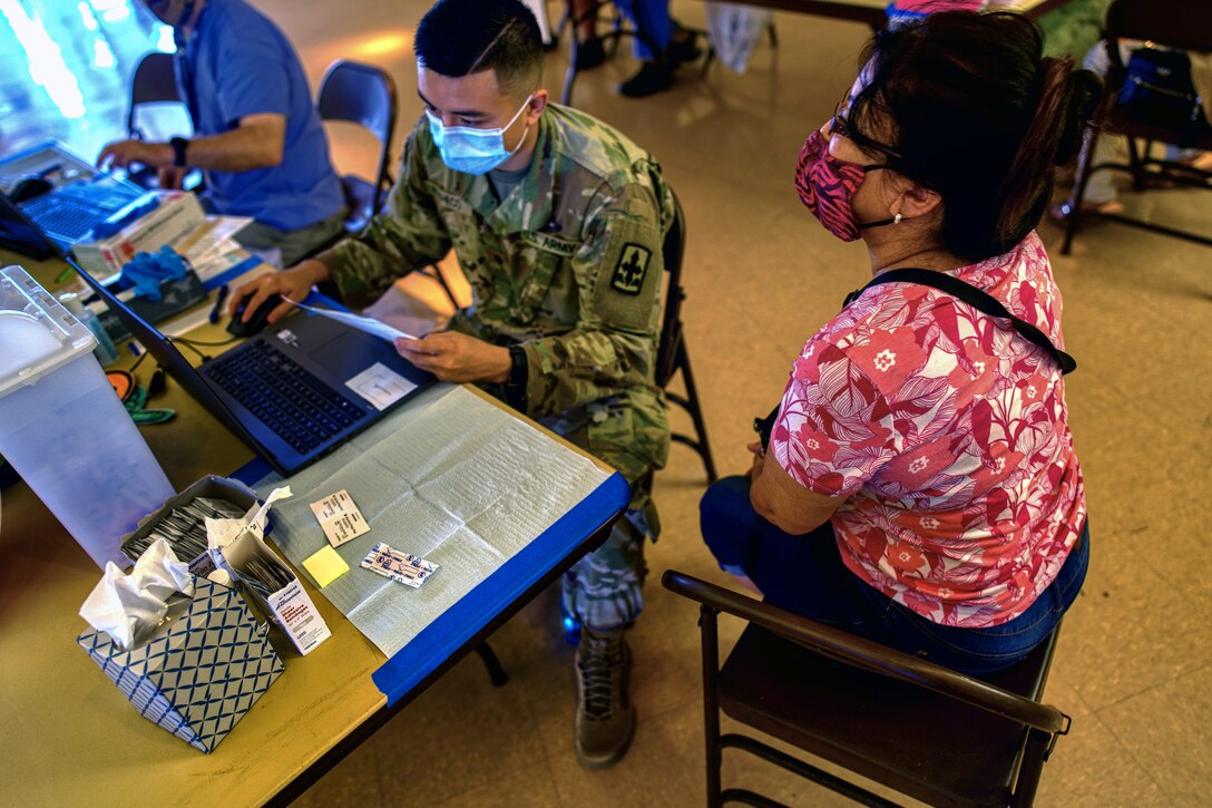 A soldier wearing a face mask sits at a table and checks in a resident on his laptop before administering a COVID-19 vaccine.