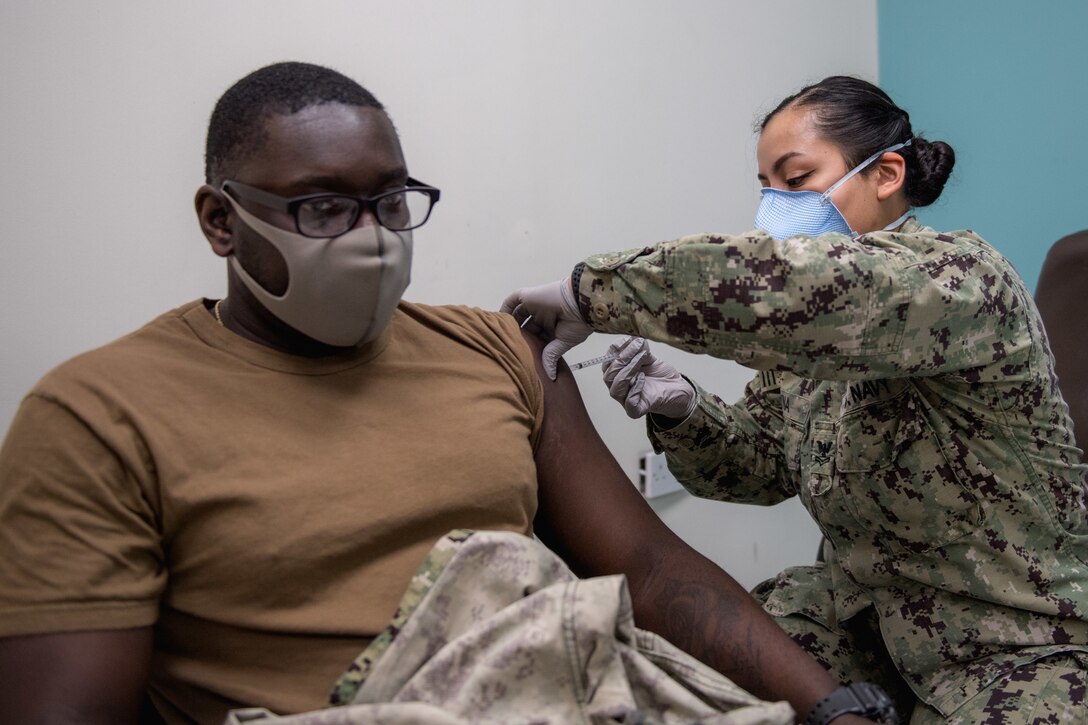 A sailor wearing a face mask and gloves administers a vaccine to another sailor.