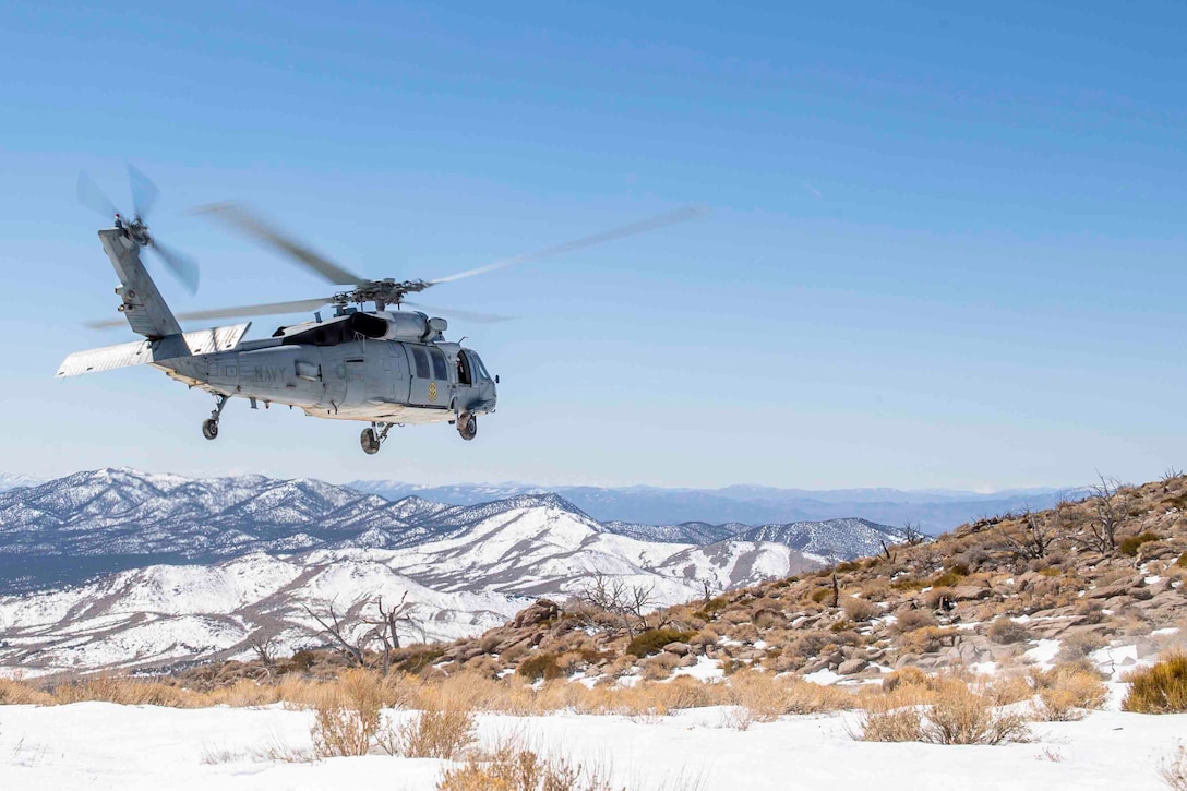 A Navy helicopter flies over a snowy terrain; a mountain is seen in the background.