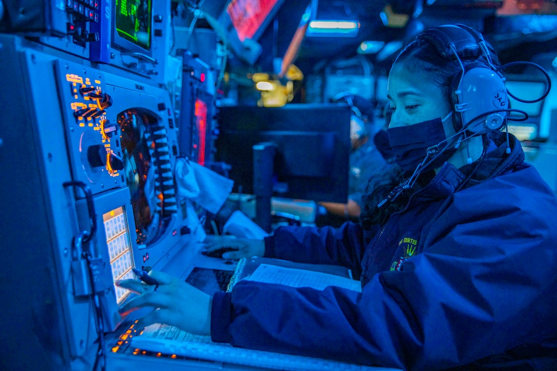 A sailor illuminated by blue light sits in front of digital monitors.