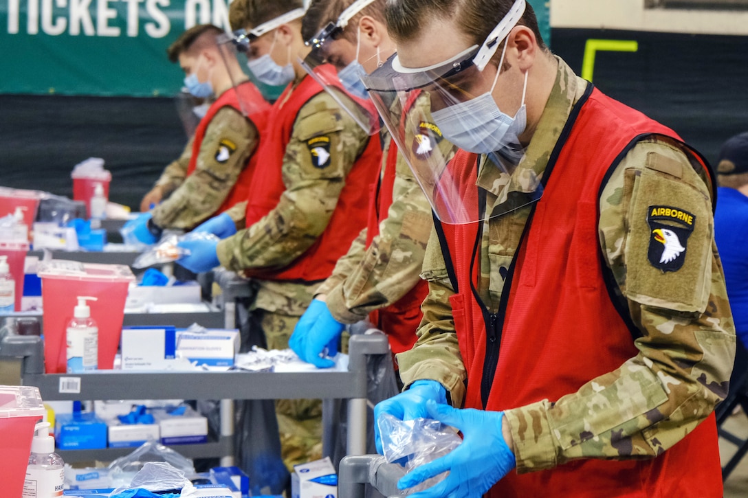 Four soldiers wearing face shields, masks and gloves stand at small tables filled with medical supplies.