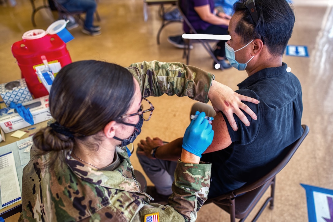 A female soldier wearing a face mask and one glove administers an injection to a man wearing a face mask.
