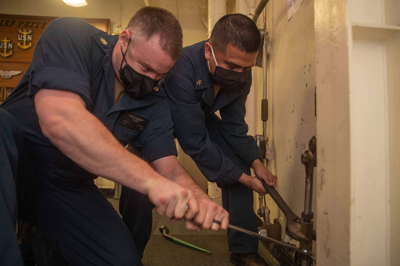 Two men in uniforms use wrenches to turn a pipe.