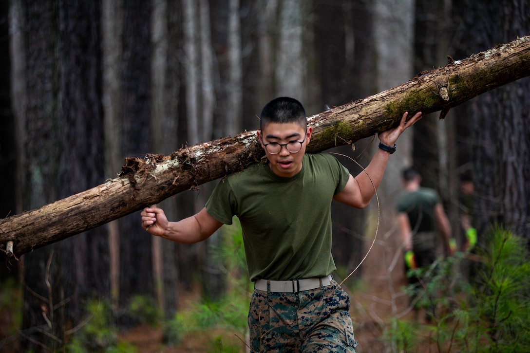A Marine carries a log on his back while walking through a forest.