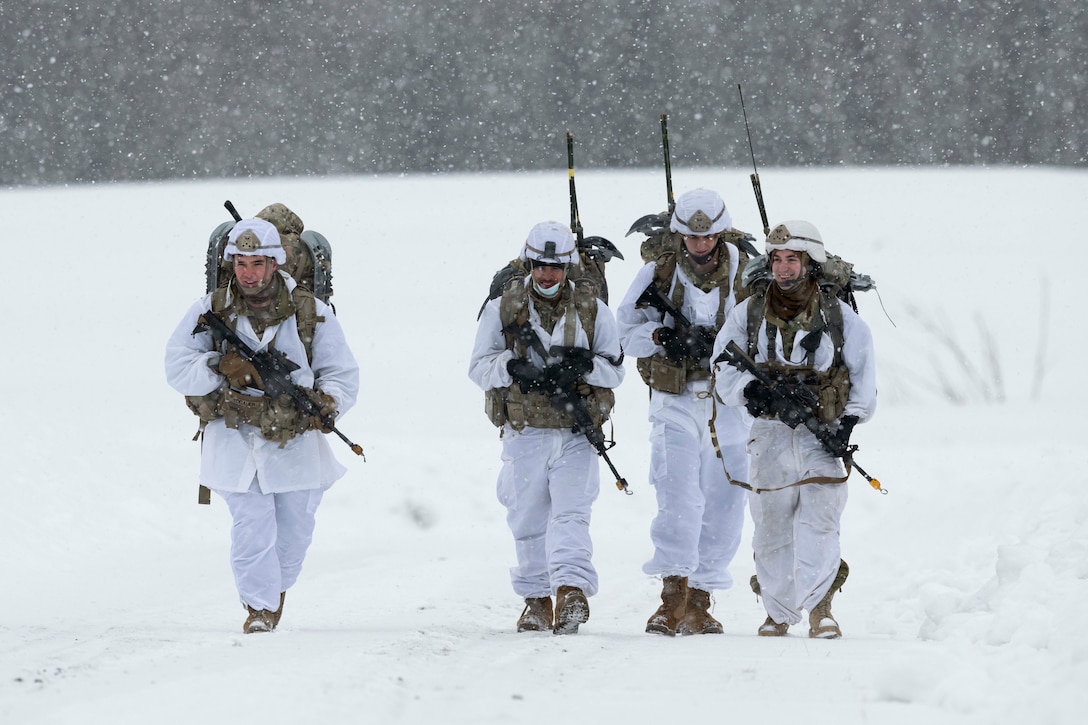 Four soldiers carrying weapons and dressed in winter gear walk through the snow.