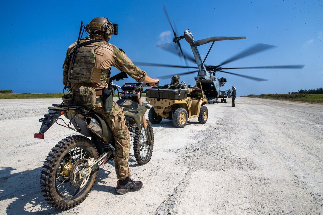 Service members wait in a small vehicle and on a motorcycle  to board a large helicopter.