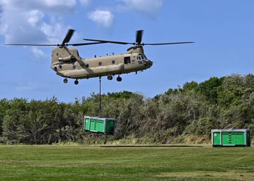 A helicopter carries a container from a rope over grass.