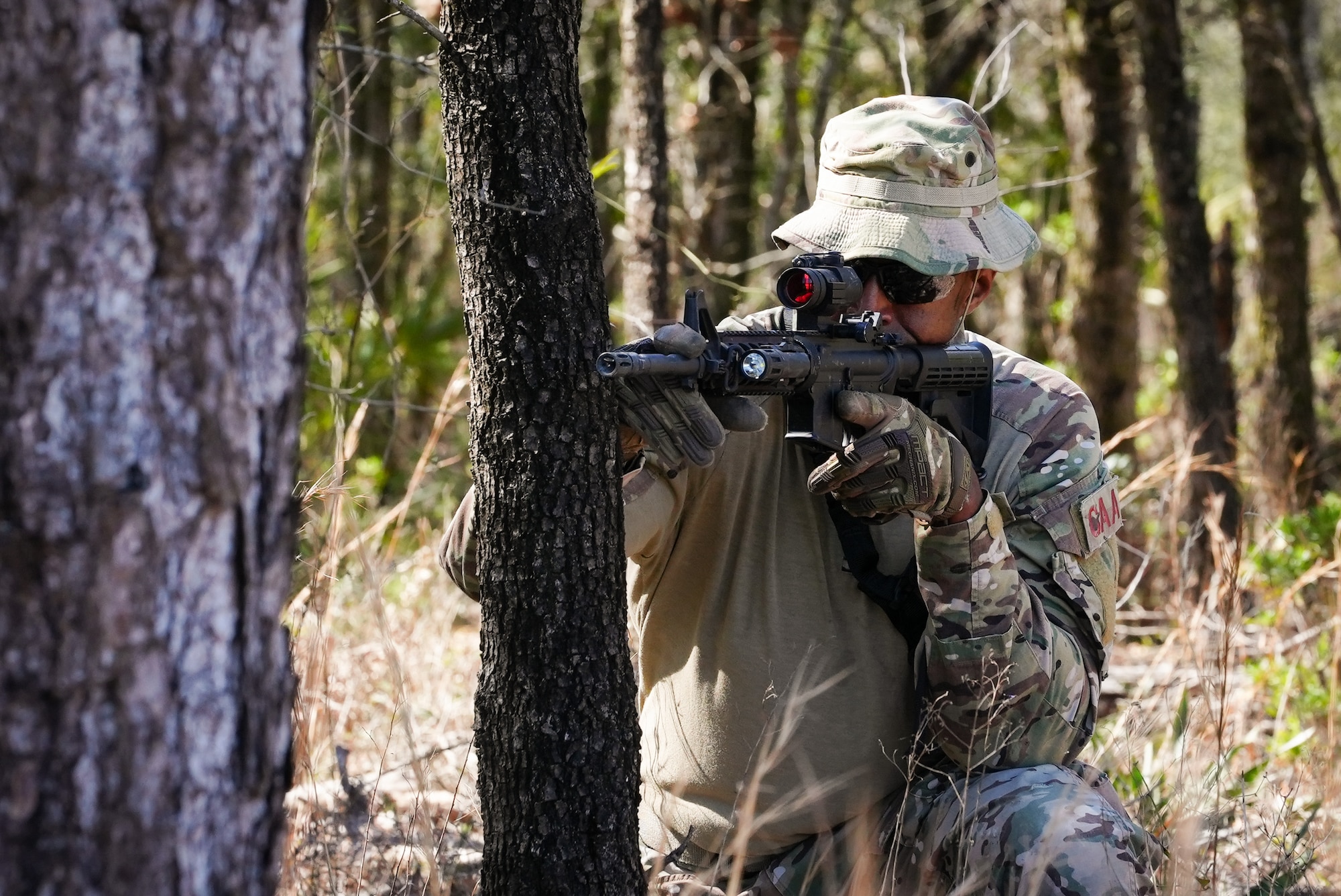 A kneeling Airman aims a weapon around a tree.