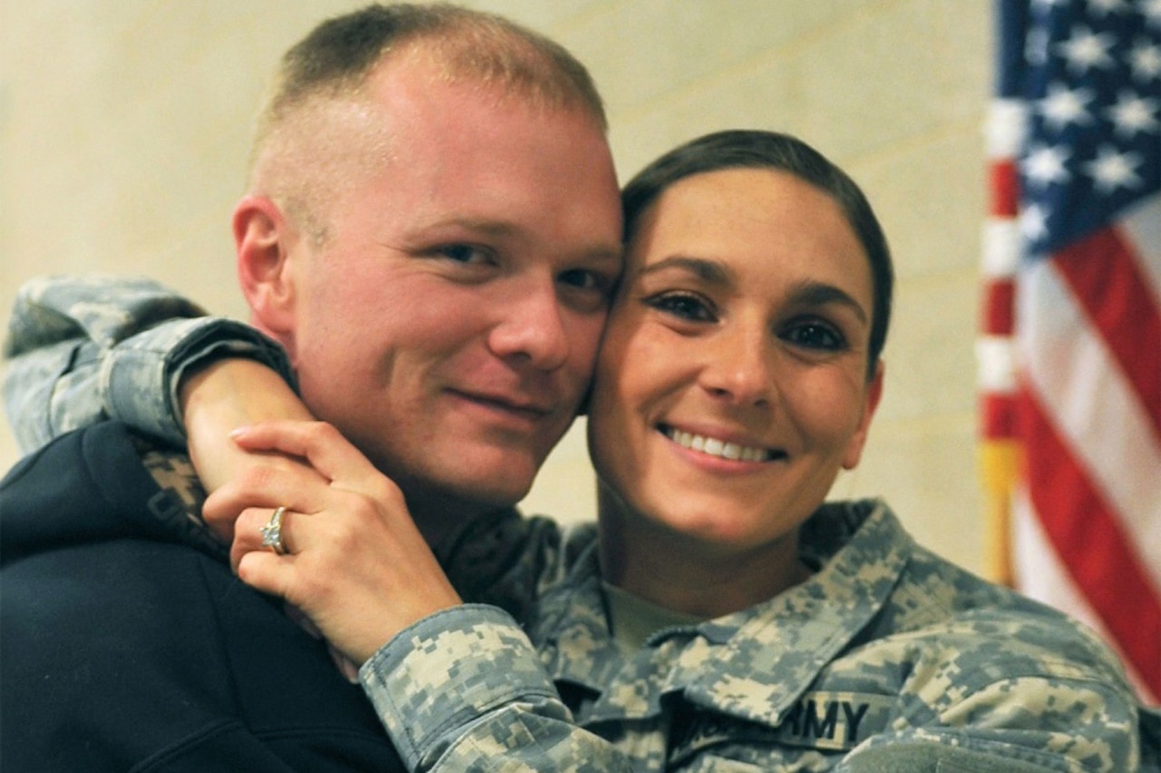 A woman in a military uniform puts her arms around the neck of a man.