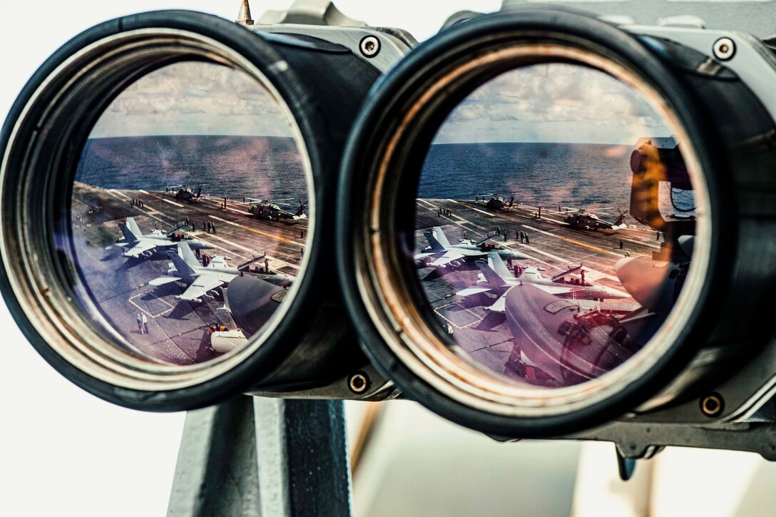 Sailors work on the flight deck of a ship next to aircraft as seen through binoculars.