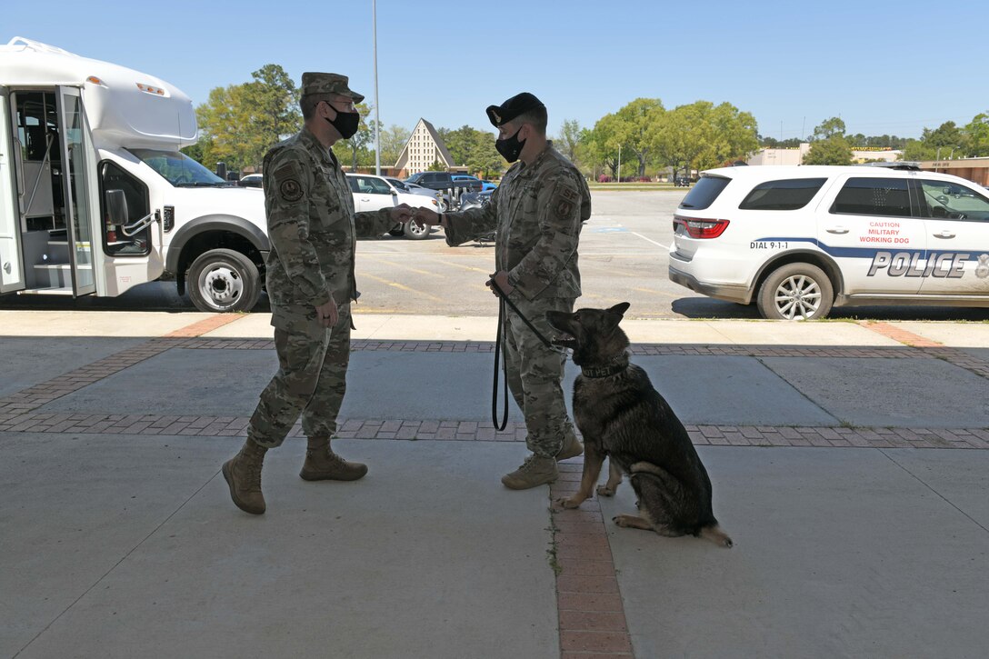 Photo shows Chief handing his coin to an Airman while his Military Working Dog sits at his feet.
