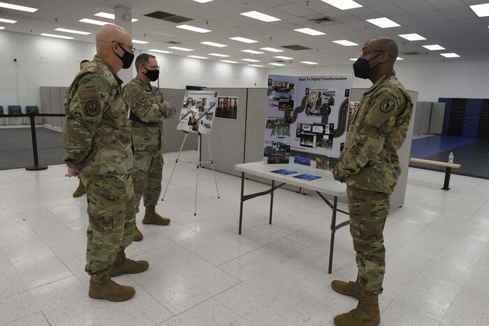 Photo shows three men looking at a board on an easel.