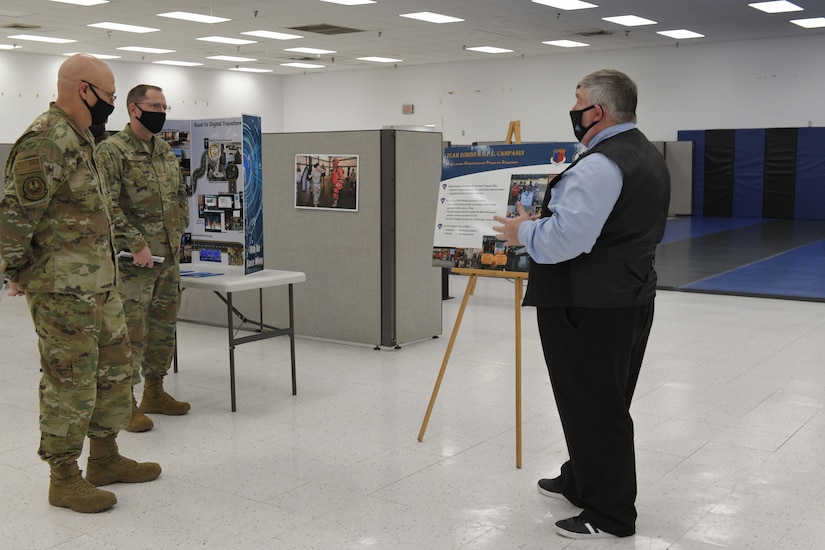 Photo shows three men looking at a poster board on an easel.