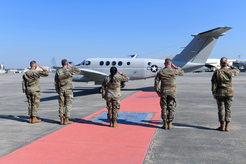 Photo  shows five people standing in a line saluting an aircraft on the flight line.