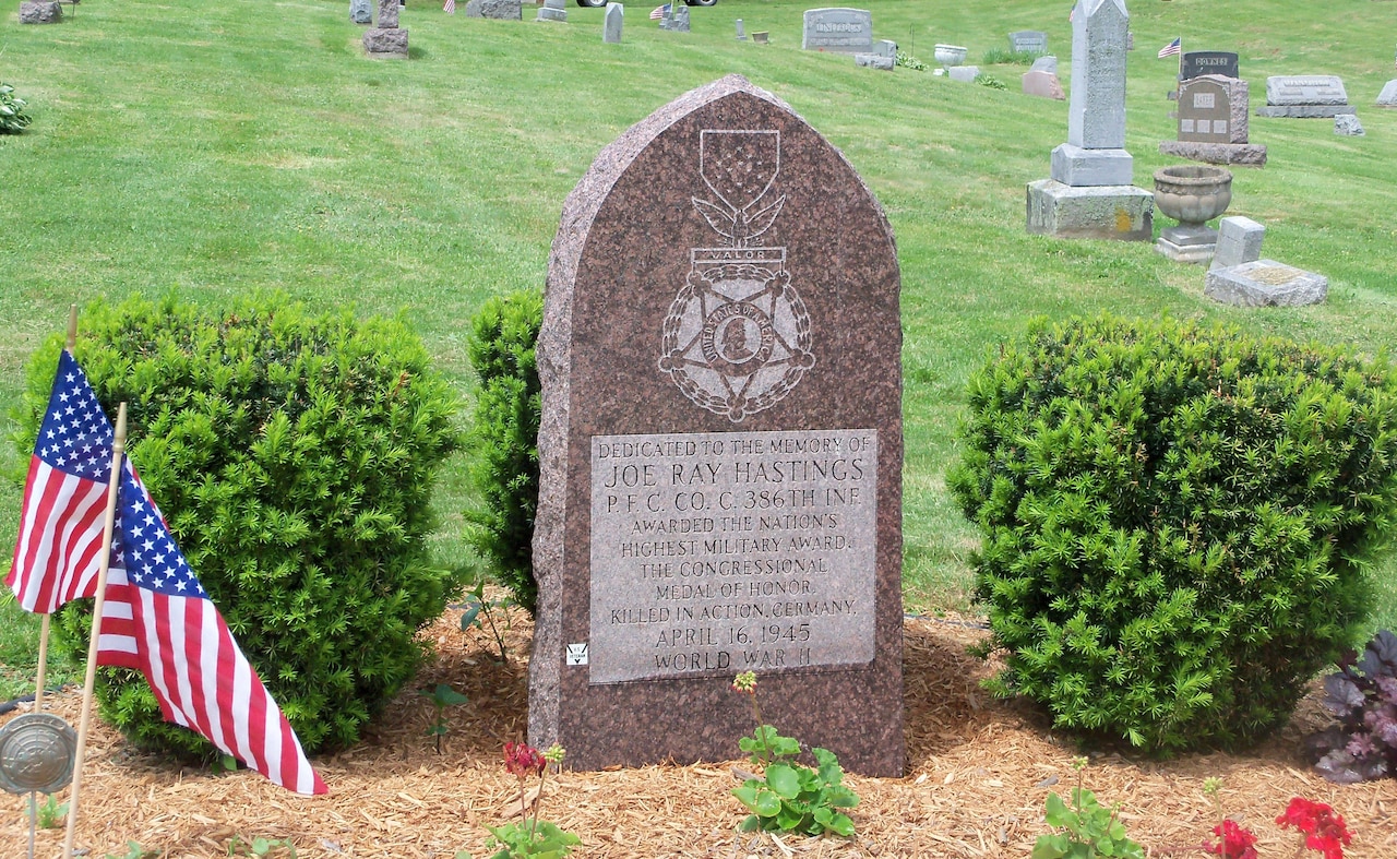 A gravestone, three small bushes and two American flags adorn a grave.