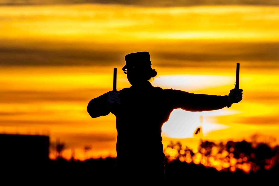 An airman shown in silhouette signals while holding onto two objects.