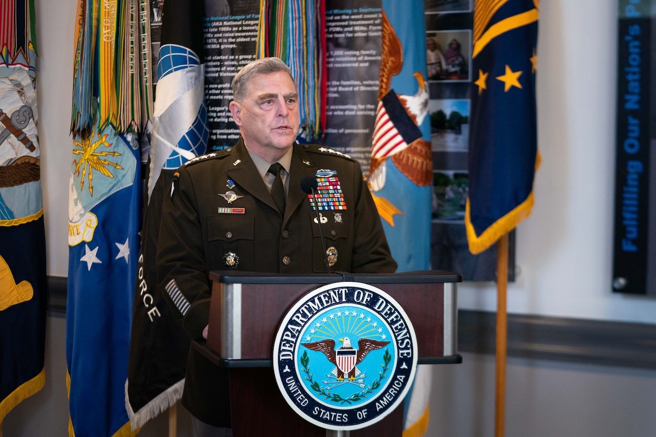A general officer, standing at a lectern with flags behind him, speaks.