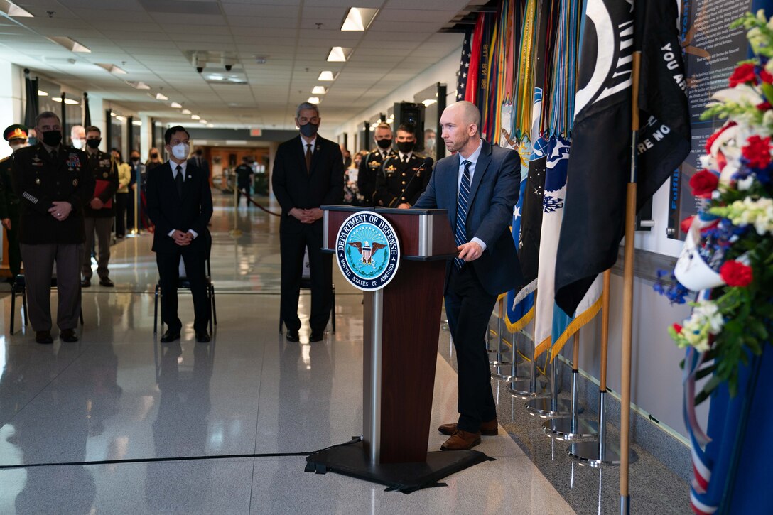 A civilian stands at a lectern with flags behind him and speaks to a group at a commemoration ceremony.