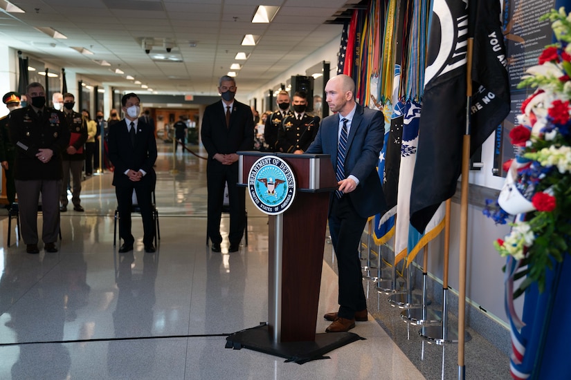 A civilian stands at a lectern with flags behind him and speaks to a group at a commemoration ceremony.