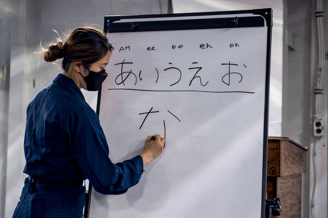 A sailor standing in a room writes Japanese characters on a whiteboard.