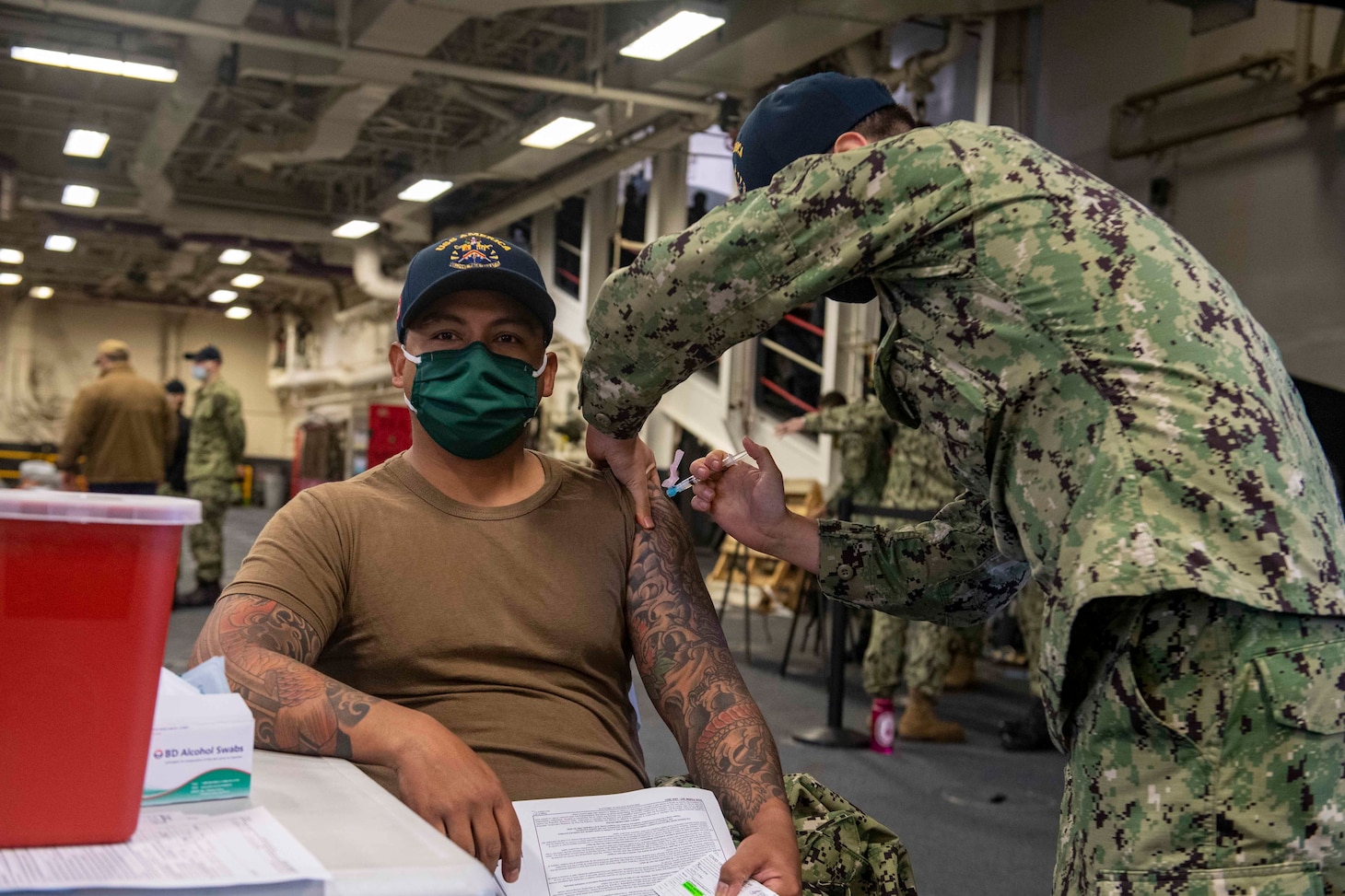 SASEBO, Japan (Feb. 26, 2021) Aviation Boatswain’s Mate (Fuel) 1st Class Michael Caballero, from Humble, Texas, assigned to the forward-deployed amphibious assault ship USS America (LHA 6), receives the first dose of the Moderna COVID-19 vaccine in the ship’s vehicle stowage area. America, lead ship of the America Amphibious Ready Group, along with the 31st Marine Expeditionary Unit, is operating in the U.S. 7th Fleet area of responsibility to enhance interoperability with allies and partners and serve as a ready response force to defend peace and stability in the Indo-Pacific region. (U.S. Navy photo by Mass Communication Specialist Seaman Matthew Cavenaile)