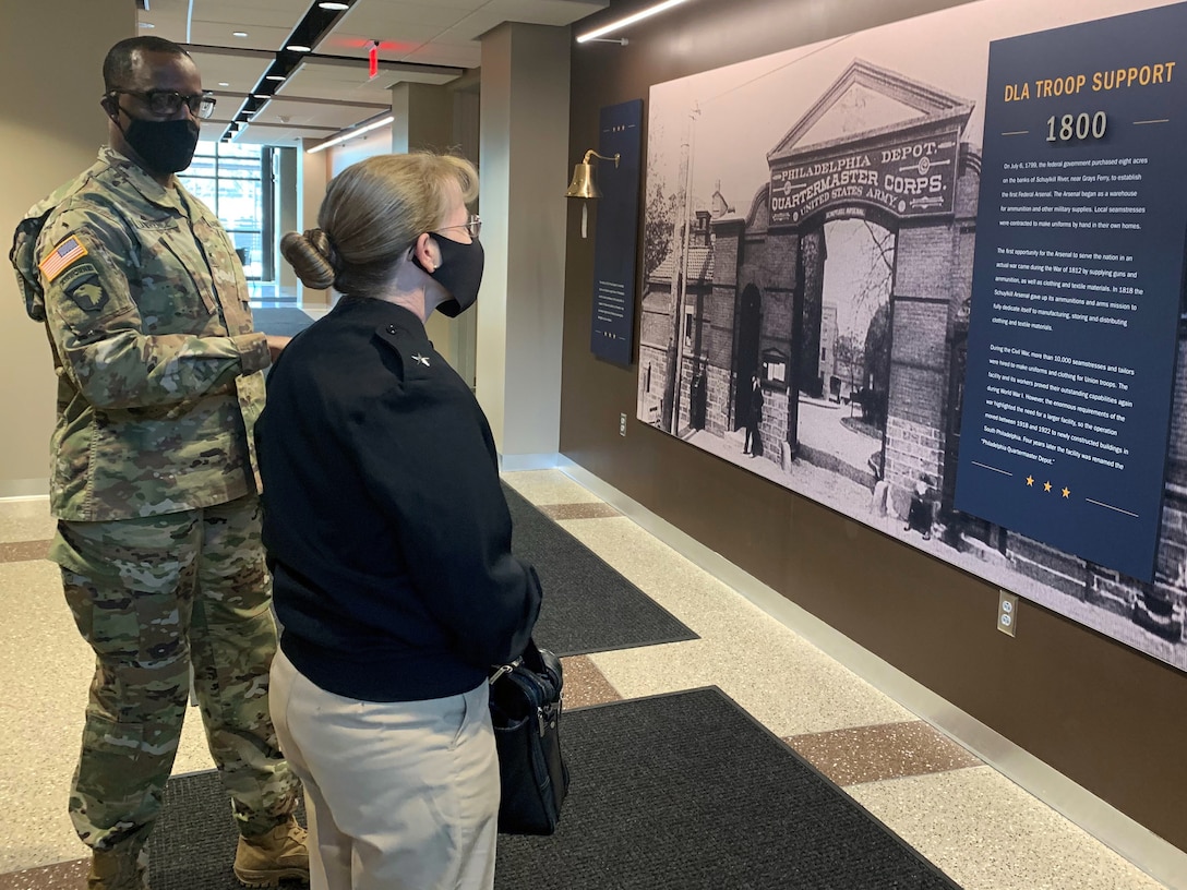 A man in a military uniform speaks to a woman in uniform while standing in front of an historical mural with summary text about the history of DLA Troop Support in Philadelphia.