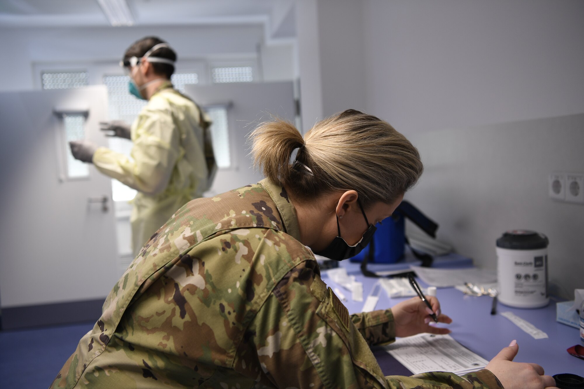 U.S. Air Force Senior Airman Kristen Nicola, 52nd Medical Group Public Health technician (front), prepares Covid test paperwork as U.S. Air Force Staff Sgt. Paul Hukreide, 52nd Medical Group Warriors Operational Health Clinic NCO in charge, prepares to administer a Covid test at Spangdahlem Air Base Germany, April 1, 2021. For Covid testing, some of the duties Public Health is responsible for is interviewing, screening, and contact tracing patients. (U.S. Air Force photo by Tech. Sgt. Warren D. Spearman Jr.)