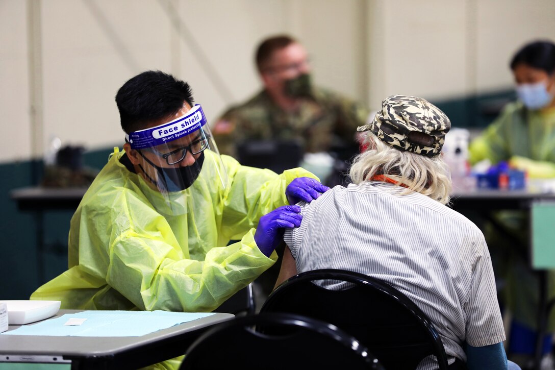 A soldier wearing personal protective equipment pushes up the sleeve of an elderly person, who's seated in a chair next to a small table.