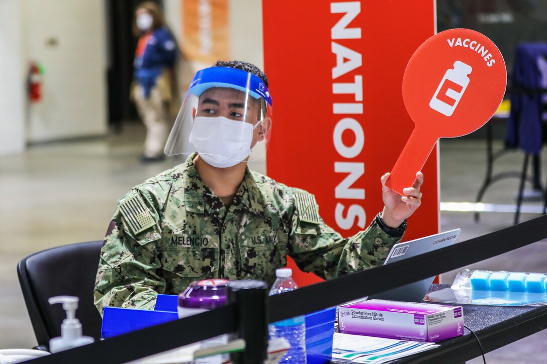 A soldier wearing a face mask and shield sits at a table holding medical supplies and holds up a sign bearing the word "vaccines."