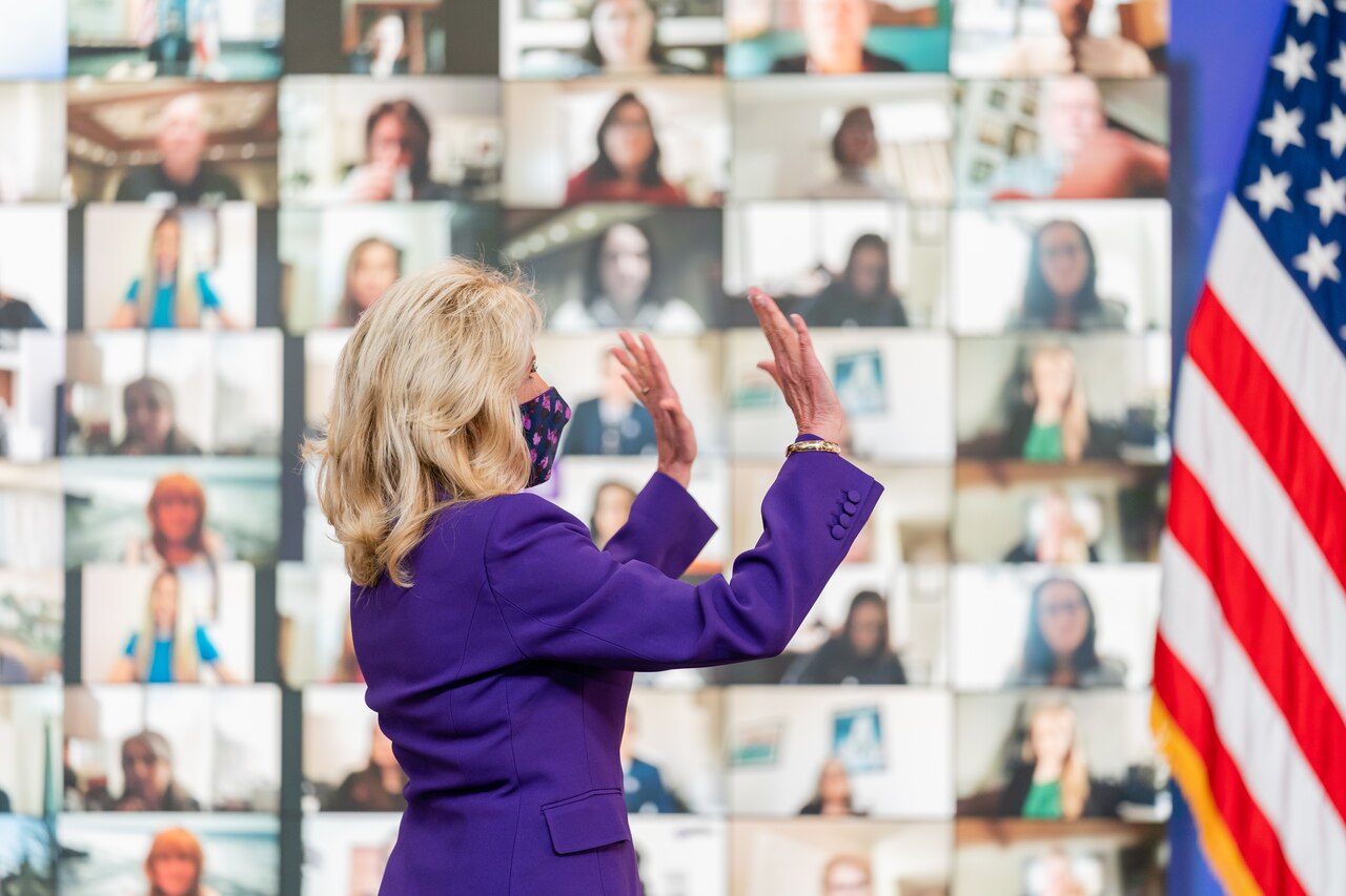 A woman in a suit, with her back to the camera, holds up both hands to the monitors that were behind her showing the faces of those virtually attending the event. An American flag is to her right.