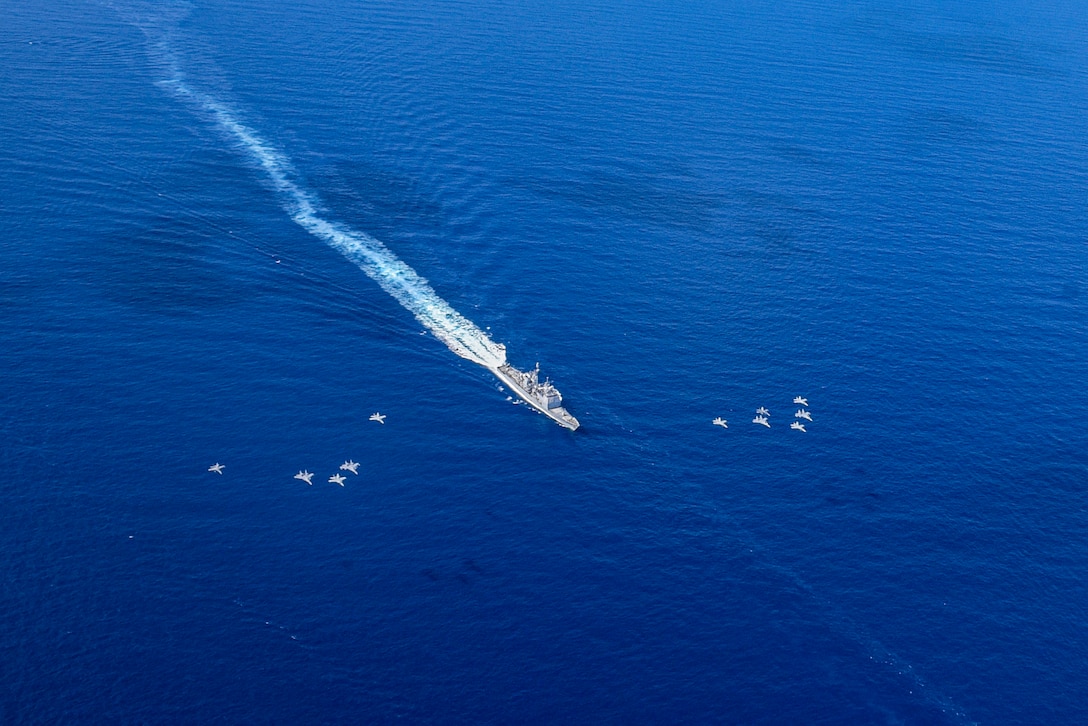 A ship, scene from above, leaves a white wake as it travels in vivid blue waters, with aircraft flying in formation on either side..