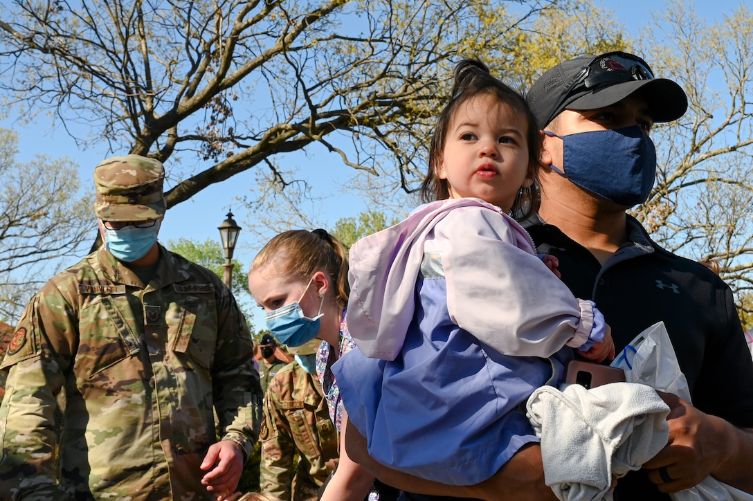 An adult holds a child outside as service members gather behind them.