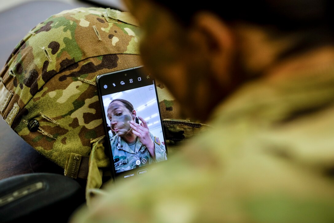 A soldier looks at her reflection in a cell phone while applying face paint.