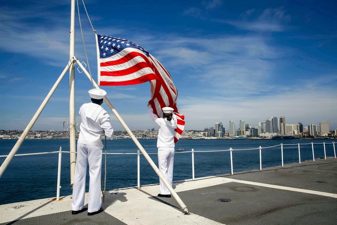 Two sailors handle a flag on a ship's flight deck, with a city skyline in the distance.