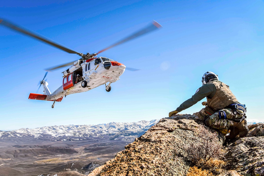 A sailor sits on a rock while a helicopter hovers above.