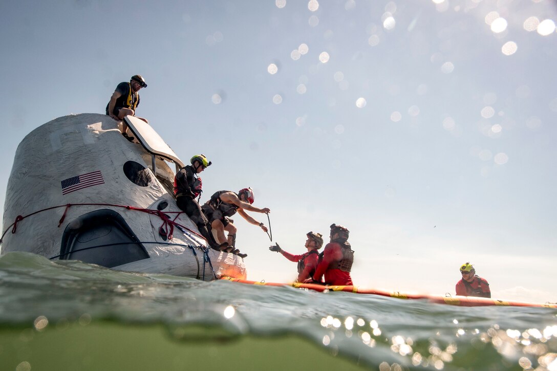 Airmen sit atop a space capsule in the water as others stand in the water nearby.