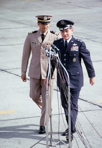 Two men in nice uniforms walk towards some microphones.