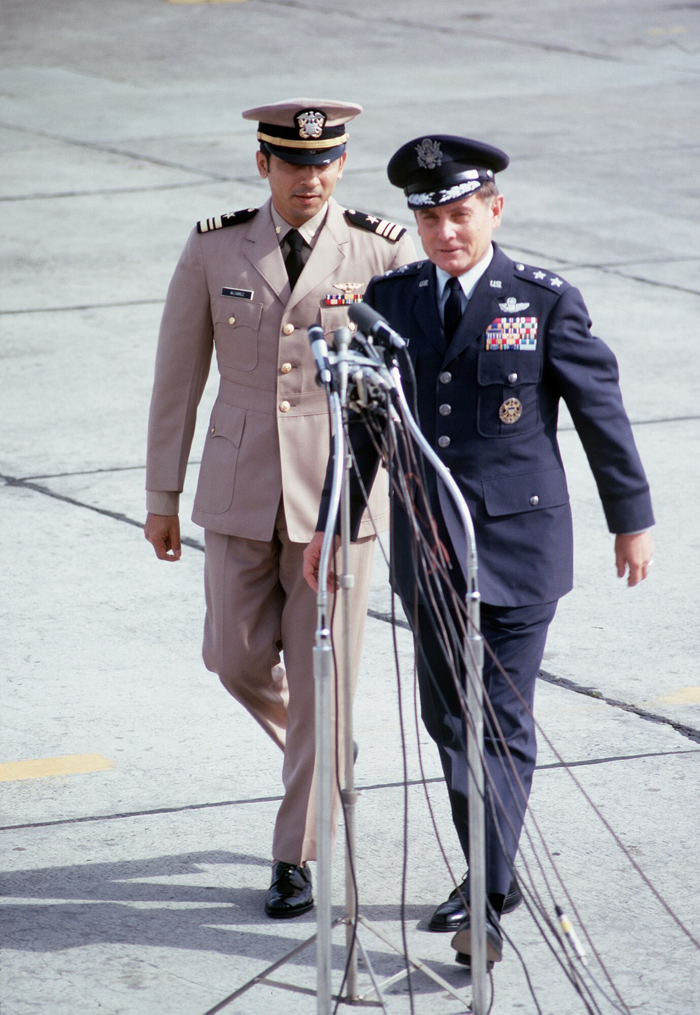 Two men in nice uniforms walk towards some microphones.