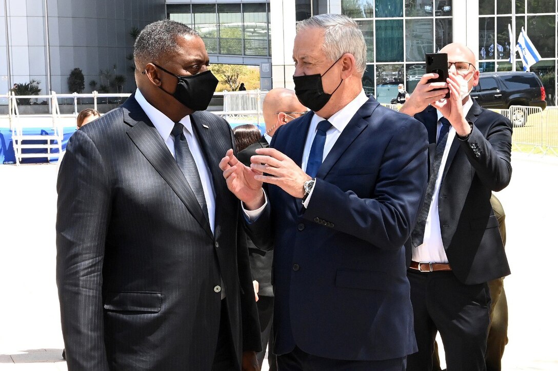 Two men wearing masks speak to each other while a third man takes a picture.