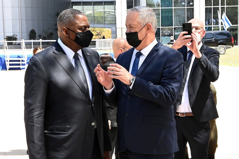 Two men wearing masks speak to each other while a third man takes a picture.