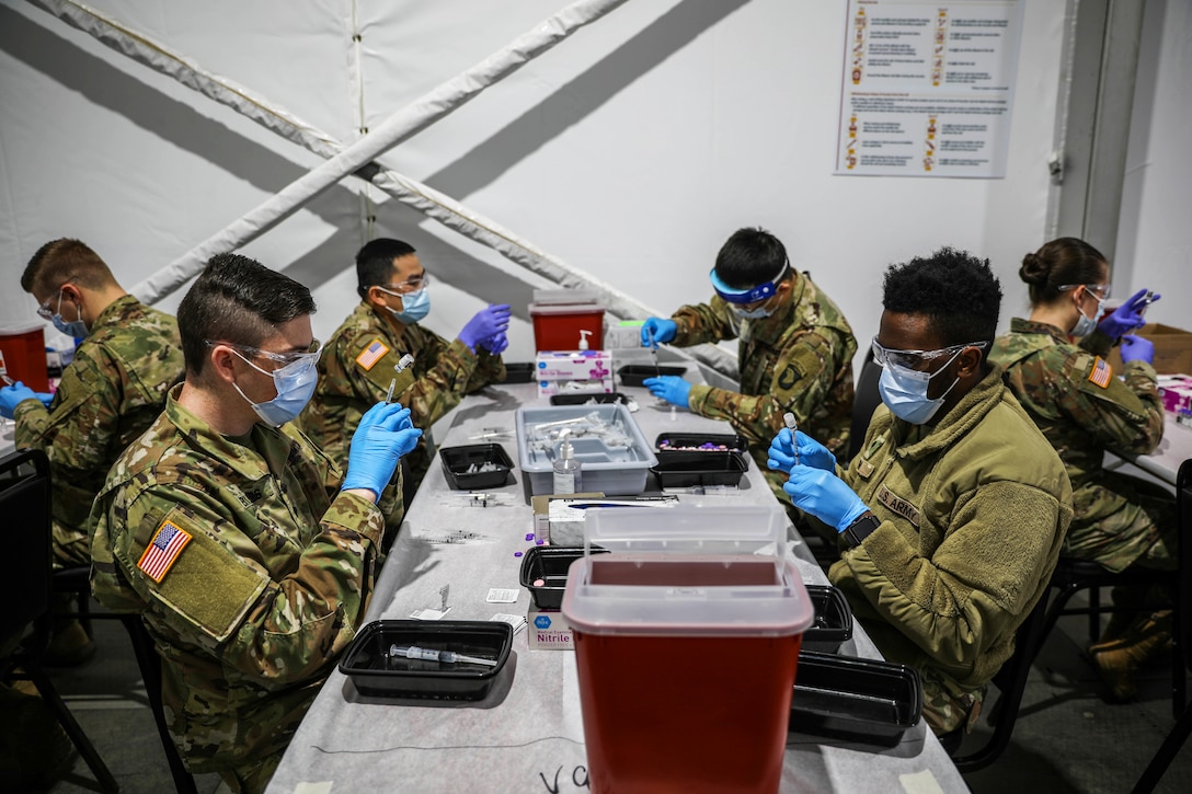 Several soldiers wearing face masks and gloves seated at a table on both sides as well as two more tables on the right and left prepare new batches of the COVID-19 vaccines.