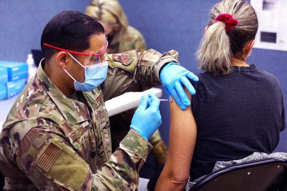 An airman wearing a face mask and gloves kneels down to give an injection to a woman, who's seated in a chair.