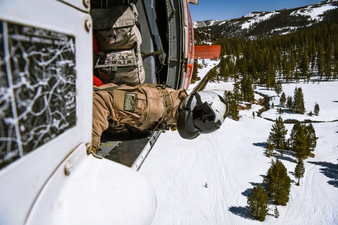 A man leans out of a helicopter as it flies above a snowy mountain.