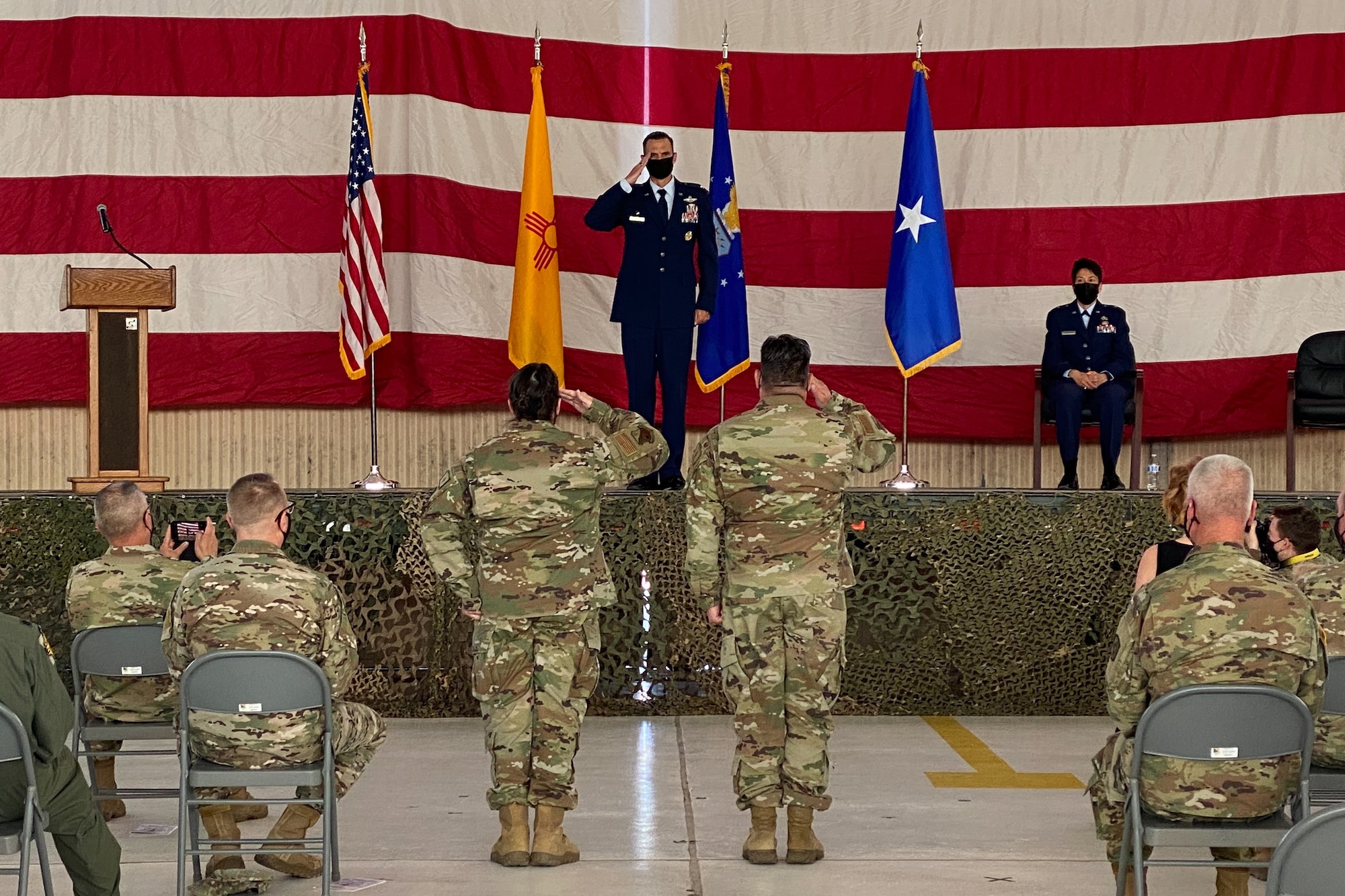 Col. Sefzik stands on the center of the stage holding a salute, with an American flag, New Mexico State flag, Air National Guard flag, and brigadier general one star flag behind him. on the floor two airmen salute Col. Sefzik as a crowd of soldiers and airmen watch