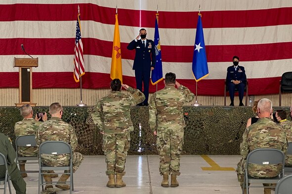 Col. Sefzik stands on the center of the stage holding a salute, with an American flag, New Mexico State flag, Air National Guard flag, and brigadier general one star flag behind him. on the floor two airmen salute Col. Sefzik as a crowd of soldiers and airmen watch