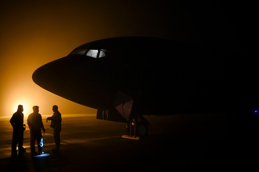 Airmen stand near an aircraft under a sunlit sky as shown in silhouette.